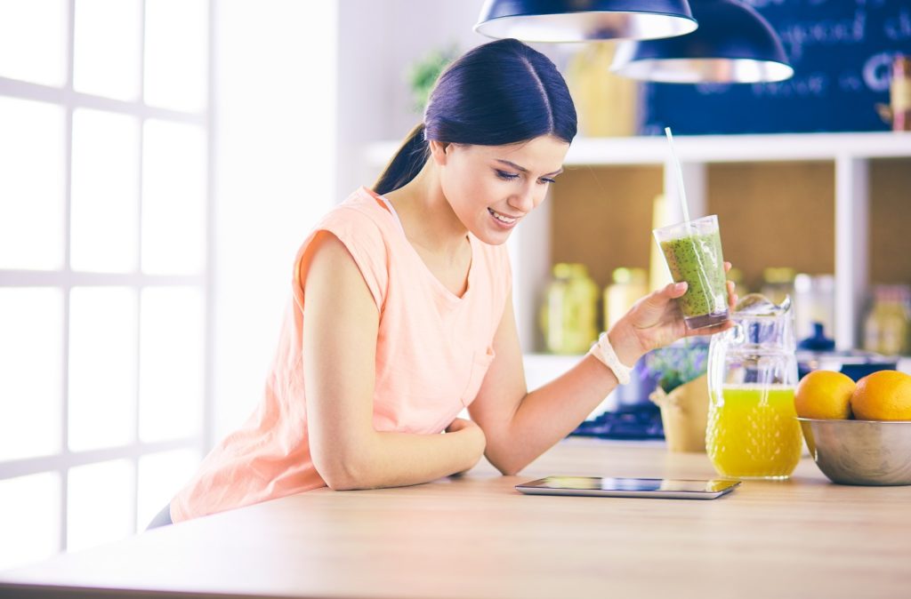 mulher tomando suco verde na cozinha, olhando para um tablet