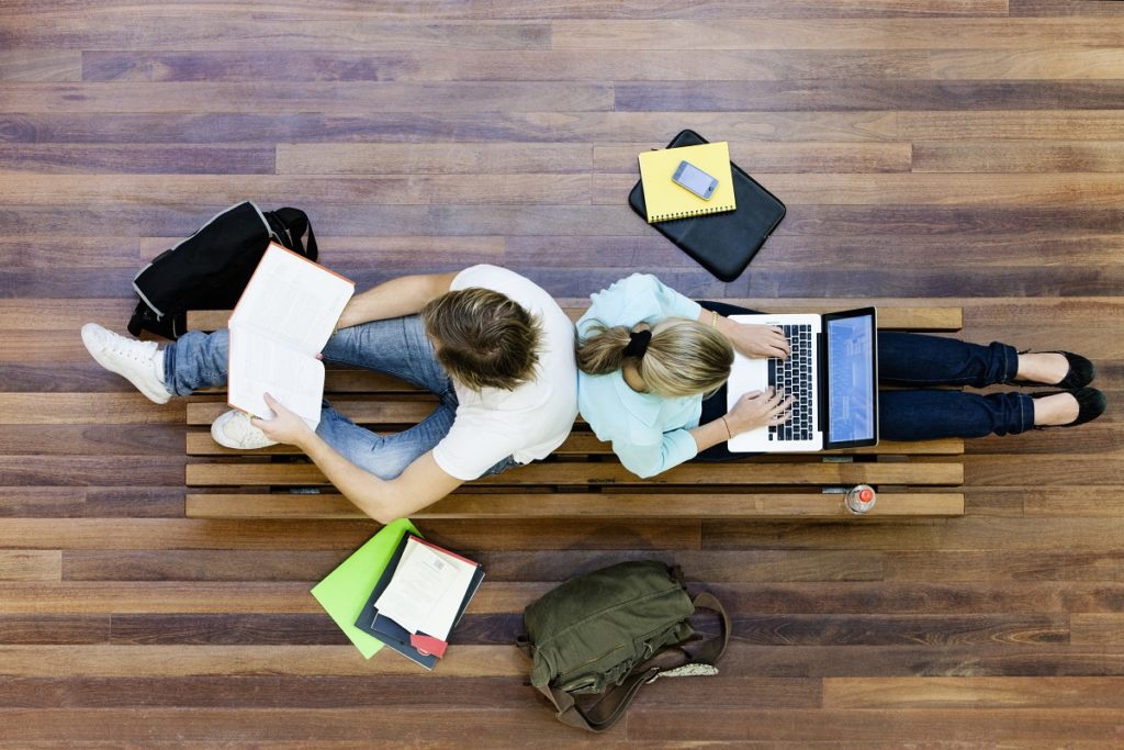 duas pessoas sentadas de costas uma para a outra, estudando em um banco de madeira