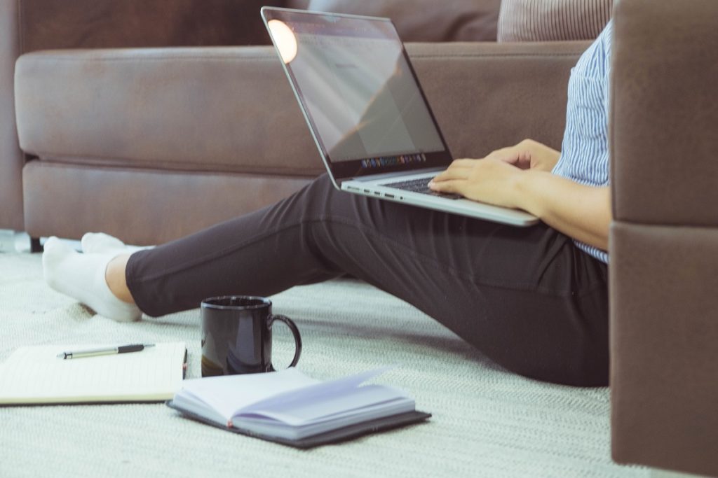 pessoa sentada no tapete com notebook no colo, com cadernos e caneca ao lado estudando para concurso vestibular