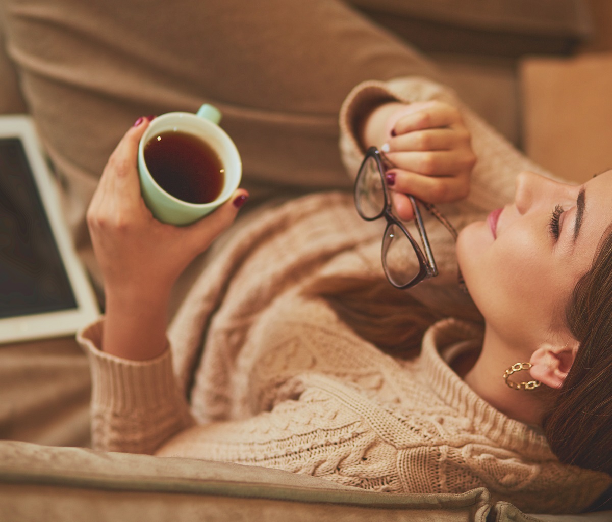 mulher vestindo tricô bege, relaxando com uma xícara de chá e segurando um par de óculos